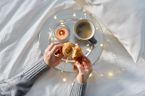 food, breakfast and people concept - hands of woman eating croissant with coffee in bed