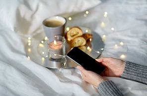 technology, hygge and christmas concept - hands of woman with smartphone, croissant and coffee in bed