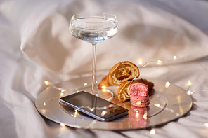 hygge and lifestyle concept - glass of champagne wine, smartphone, croissants, macaroons and garland lights in bed at home