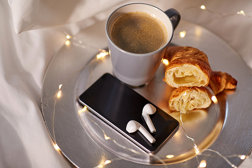 morning, hygge and breakfast concept - smartphone, wireless earphones, cup of coffee and croissants with garland lights on plate in bed at home