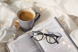 morning, hygge and breakfast concept - cup of coffee, book, glasses and garland lights in bed at home