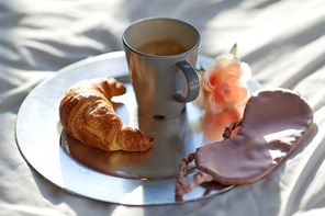 morning, hygge and breakfast concept - croissant, cup of coffee and eye sleeping mask on plate in bed at home