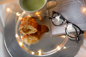 morning, hygge and breakfast concept - croissants, cup of matcha tea book and glasses in bed at home