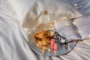 hygge and lifestyle concept - glass of champagne wine, croissants, macaroons with book and glasses in bed at home