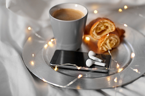 morning, hygge and breakfast concept - smartphone, wireless earphones, cup of coffee and croissants with garland lights on plate in bed at home