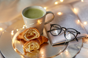 hygge and breakfast concept - croissants, cup of matcha tea book and glasses in bed at home
