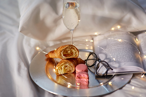 hygge and lifestyle concept - glass of champagne wine, croissants, macaroons with book and glasses in bed at home