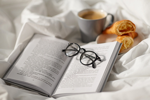 morning, hygge and breakfast concept - croissants, cup of coffee, book and glasses in bed at home