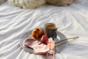morning, hygge and breakfast concept - croissant, cup of coffee, macaroons and eye sleeping mask on plate in bed at home