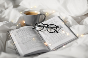 morning, hygge and breakfast concept - cup of coffee, book, glasses and garland lights in bed at home