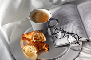 morning, hygge and breakfast concept - croissants, cup of coffee, book and glasses in bed at home