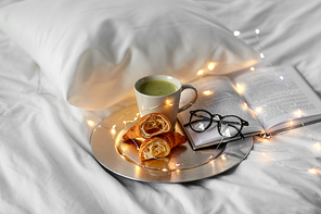 morning, hygge and breakfast concept - croissants, cup of matcha tea book and glasses in bed at home