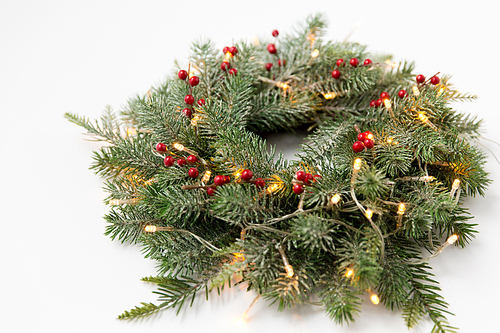 winter holidays, new year and decorations concept - wreath of fir branches with red berries and garland lights on white background