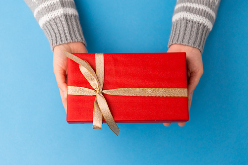 holiday, presents and greetings concept - hands holding small christmas red gift box with golden bow on blue background
