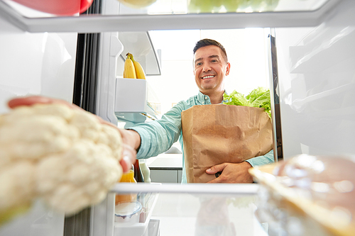 eating, diet and storage concept - smiling middle-aged man with new purchased food in paper bag putting cauliflower to fridge at home
