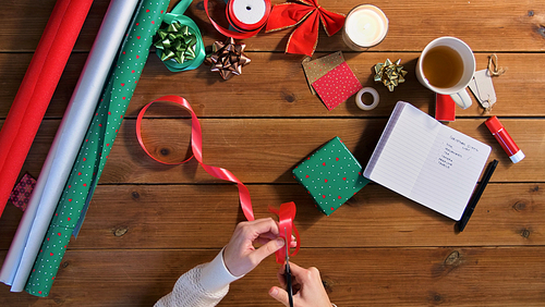 christmas, packing and holidays concept - hands with scissors cutting red ribbon for gift box on wooden table