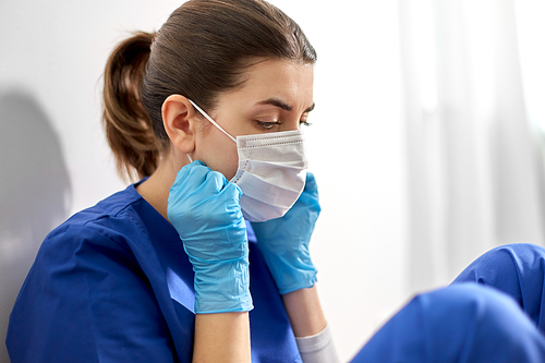 medicine, healthcare and pandemic concept - sad young female doctor or nurse wearing face protective mask for protection from virus disease sitting on floor at hospital