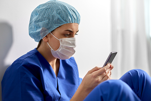 medicine, healthcare and pandemic concept - sad young female doctor or nurse with smartphone wearing face protective mask for protection from virus disease sitting on floor at hospital