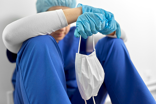 medicine, healthcare and pandemic concept - sad young female doctor or nurse holding face protective mask for protection from virus disease sitting on floor at hospital