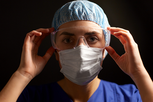 health, medicine and pandemic concept - close up of young female doctor or nurse wearing goggles, hat and face protective mask for protection from virus disease over black background