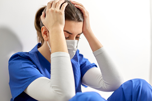 medicine, healthcare and pandemic concept - sad young female doctor or nurse wearing face protective mask for protection from virus disease sitting on floor and holding to head