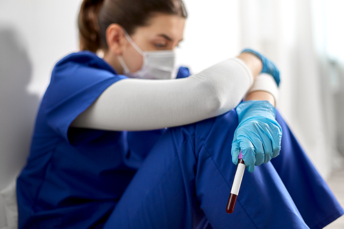 medicine, healthcare and pandemic concept - sad young female doctor or nurse sitting on floor at hospital and holding beaker with blood test