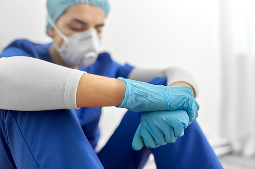 medicine, healthcare and pandemic concept - sad young female doctor or nurse wearing face protective mask or respirator for protection from virus disease sitting on floor at hospital