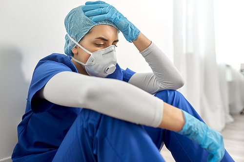 medicine, healthcare and pandemic concept - sad young female doctor or nurse wearing face protective mask or respirator for protection from virus disease holding to head