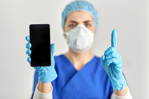 medicine, health and pandemic concept - close up of young female doctor or nurse wearing face protective mask or respirator for protection from virus disease with smartphone pointing finger up