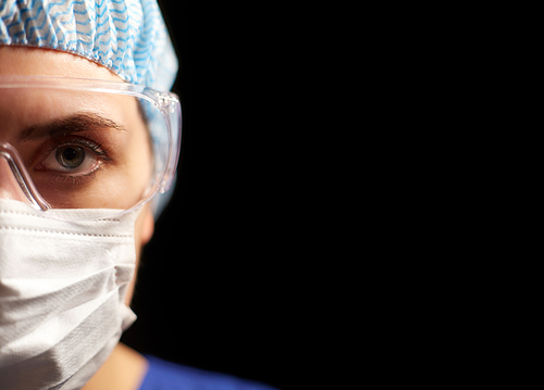 health, medicine and pandemic concept - close up of young female doctor or nurse wearing goggles, hat and face protective mask for protection from virus disease over black background