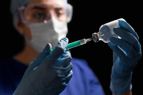 health, medicine and pandemic concept - female doctor or nurse wearing goggles, glove and face protective mask with syringe and drug in glass jar over black background