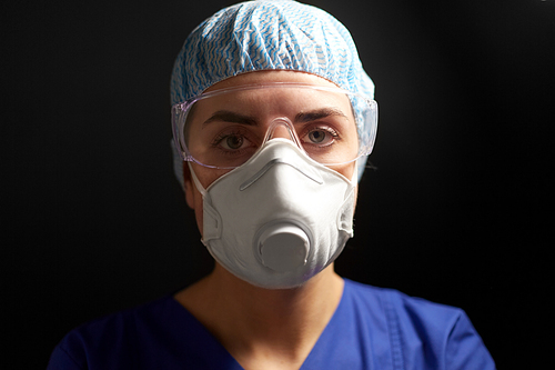 medicine, health and pandemic concept - close up of young female doctor or nurse wearing goggles, hat and face protective mask or respirator for protection from virus disease over black background