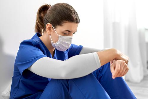 medicine, healthcare and pandemic concept - sad young female doctor or nurse wearing face protective mask for protection from virus disease sitting on floor at hospital