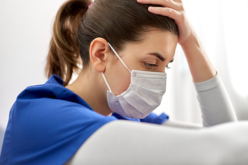 medicine, healthcare and pandemic concept - sad young female doctor or nurse wearing face protective mask for protection from virus disease sitting on floor and holding to head