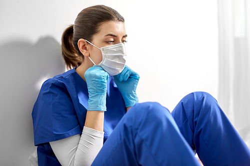 medicine, healthcare and pandemic concept - sad young female doctor or nurse wearing face protective mask for protection from virus disease sitting on floor at hospital