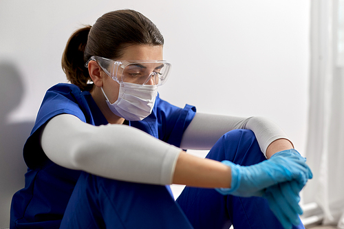 medicine, healthcare and pandemic concept - sad young female doctor or nurse wearing goggles and face protective mask for protection from virus disease sitting on floor at hospital