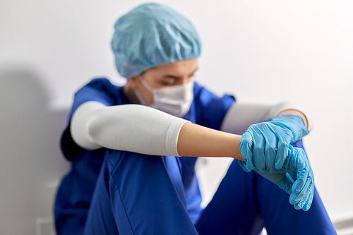 medicine, healthcare and pandemic concept - sad young female doctor or nurse wearing face protective mask for protection from virus disease sitting on floor at hospital