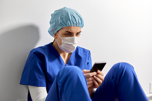 medicine, healthcare and pandemic concept - sad young female doctor or nurse with smartphone wearing face protective mask for protection from virus disease sitting on floor at hospital