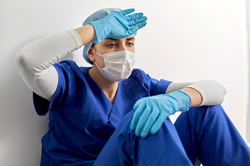 medicine, healthcare and pandemic concept - sad young female doctor or nurse wearing face protective mask for protection from virus disease sitting on floor at hospital