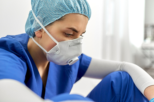 medicine, healthcare and pandemic concept - sad young female doctor or nurse wearing face protective mask or respirator for protection from virus disease sitting on floor at hospital