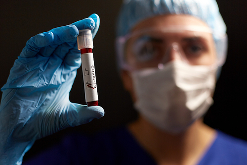 medicine, health and pandemic concept - close up of young female doctor or nurse wearing goggles, glove and face protective mask holding beaker with coronavirus blood test over black background