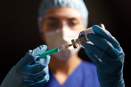 health, medicine and pandemic concept - female doctor or nurse wearing goggles, glove and face protective mask with syringe and drug in glass jar over black background