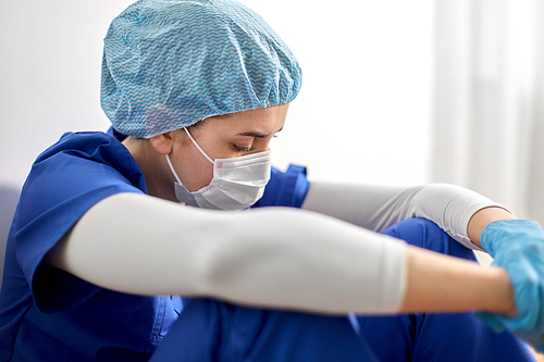 medicine, healthcare and pandemic concept - sad young female doctor or nurse wearing face protective mask for protection from virus disease sitting on floor at hospital