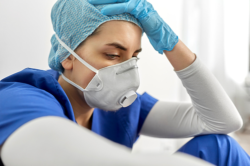 medicine, healthcare and pandemic concept - sad young female doctor or nurse wearing face protective mask or respirator for protection from virus disease holding to head