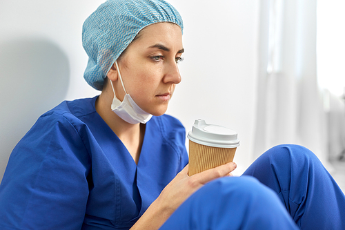 medicine, healthcare and pandemic concept - close up of sad young female doctor or nurse with disposable cup of takeaway coffee sitting on floor at hospital