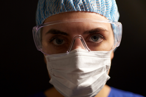 health, medicine and pandemic concept - close up of young female doctor or nurse wearing goggles, hat and face protective mask for protection from virus disease over black background