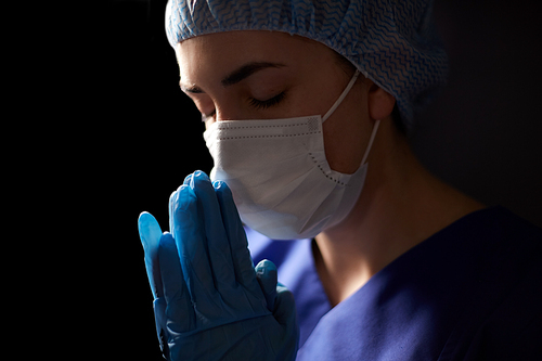 health, medicine and pandemic concept - sad young female doctor or nurse wearing face protective mask for protection from virus disease praying over black background