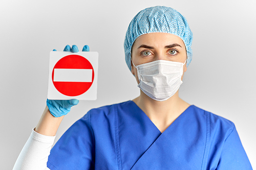 health, medicine and pandemic concept - young female doctor or nurse wearing face protective mask for protection from virus disease and hat showing stop sign over grey background