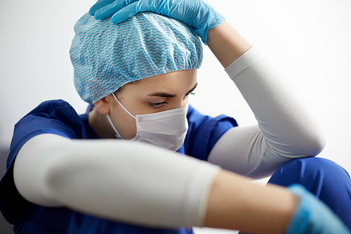 medicine, healthcare and pandemic concept - sad young female doctor or nurse wearing face protective mask for protection from virus disease holding to head