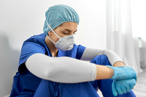 medicine, healthcare and pandemic concept - sad young female doctor or nurse wearing face protective mask or respirator for protection from virus disease sitting on floor at hospital